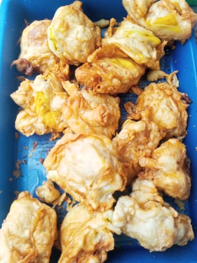 A blue tray holds a pile of golden-brown, deep-fried eggs. Some eggs reveal bright yellow yolks peeking through the crispy batter. The fried batter has an irregular, lumpy texture and various shades of golden-brown, suggesting uneven cooking or varying thickness of the batter. The eggs are clumped together, partially obscuring one another.