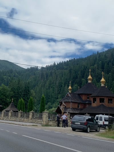 An outdoor scene in Tatariv, Ukraine, depicts a multi-domed wooden church situated against a backdrop of densely forested mountains under a partially cloudy sky. The church, constructed from dark-stained wood with dark shingled roofs, features multiple golden onion domes, each topped with a cross, indicative of Eastern Orthodox architecture. A large crucifix is mounted on the front facade of the main church structure. To the left of the main building, a smaller, similarly styled wooden structure with a single dome stands behind a stone and wood fence that borders the property. Along the fence, several tall, manicured evergreen trees are planted.
In the foreground, an asphalt road with white lane markings runs horizontally, separated from the church grounds by a gravel and dirt shoulder. Parked on this shoulder are two vehicles: a dark gray minivan (with visible license plate BY 0447 AO) positioned closer to the camera, and a white utility van parked behind it, partially obscured.
Approximately five individuals are present near the church entrance and along the fence line. One man in a light shirt and dark pants stands facing a group of others, who are loosely gathered near the church steps or along the fence. Their postures suggest they are standing and engaged in conversation. Overhead, multiple power lines traverse the upper portion of the frame against the sky. The overall environment suggests a rural or semi-rural mountainous setting.