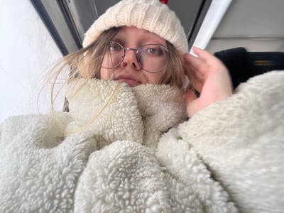 A person, likely female, is depicted in an upward-angled, close-up photograph taken within an enclosed interior space in the city of Борисово, Russia. The individual wears a cream-colored knitted beanie and a high-collared cream-colored faux shearling jacket. Round metal-rimmed glasses are present. Light-colored hair is visible around the face. The right hand is raised towards the right side of the head, partially obscuring the ear. The subject's gaze is directed at the camera. The background indicates an interior environment with a visible ceiling or upper wall section and a dark horizontal structural element. A bright exterior is subtly visible through a window on the left.