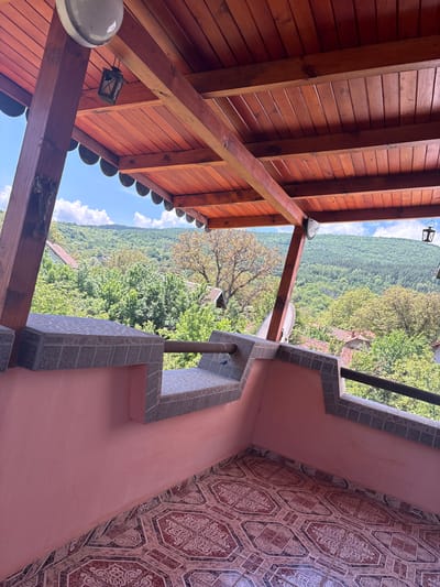 The image captures a panoramic view from a covered outdoor area, likely a balcony or porch, overlooking a vast forested valley in Svetlia, Bulgaria. The scene is illuminated by bright daylight under a clear blue sky adorned with scattered white cumulus clouds.
On the left side of the frame, a rustic wooden ceiling is prominently featured, constructed from reddish-brown planks supported by sturdy wooden beams. Two distinct light fixtures are attached to the ceiling: a modern, spherical white globe light and an older, black-framed lantern.
The right side of the image reveals a salmon-pink wall paired with an ornate tiled surface, possibly a floor or another wall section, showcasing intricate geometric and curvilinear patterns in shades of red, brown, and cream. A decorative balustrade or low wall, built from greyish-white textured tiles, forms a barrier, from which a black metal railing extends. A small satellite dish is mounted below the railing, and a water tap with associated piping is visible on the tiled structure.
The middle ground and background are dominated by a sprawling, dense green forest that covers numerous hills and valleys, stretching towards the horizon. Nestled among the trees, several houses with characteristic red or orange tiled roofs are visible, indicating a rural settlement within the natural landscape.
The overall impression is one of serene natural beauty and a peaceful rural setting. No people are visible in the image, and there is no discernible text.