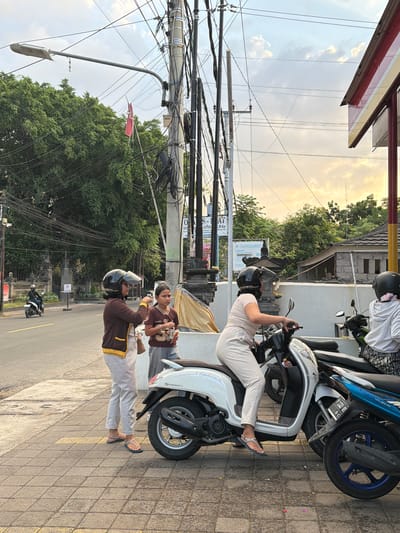 In Kubutambahan, Indonesia, three women are standing on a paved sidewalk next to a street. The woman on the far left is wearing a brown jacket with yellow trim, light grey pants, and flip-flops. She has on a black helmet with a clear visor and is holding a brown purse. Next to her, a younger girl in a brown t-shirt with the number "65" on it and grey shorts, also holds an orange object. The woman in the middle is wearing a light-colored outfit and a black helmet with a clear visor, seated on a white scooter. A second scooter, blue and black, is parked next to the white one. Behind the women, a traffic light pole is covered in tangled wires. The street is asphalt, and to the left, a person on a white scooter is visible driving away. The background consists of lush green trees, buildings with tiled roofs, and a cloudy sky at dusk, with the sun casting a warm glow.