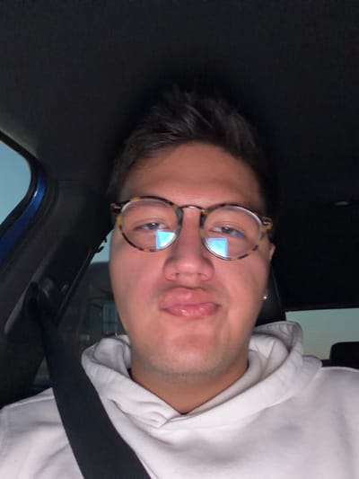 A man wearing glasses and a white hooded sweatshirt sits in the driver's seat of a vehicle, as indicated by the black seatbelt. He has dark hair and facial stubble. The car's interior is visible, including a blue panel near the window. Reflections of light are visible in his glasses. The man is looking directly at the camera with slightly pursed lips.