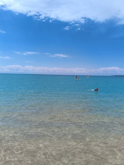 The image depicts a clear, sunny day at a beach in Bayet, Kyrgyzstan. The foreground shows shallow, clear water with visible sandy bottom. The water transitions to a deeper blue further out, stretching to the horizon. In the distance, a thin line of land is visible, possibly a distant shore or coastline. A band of white, puffy clouds is scattered across the bright blue sky.
Several individuals are engaged in water activities. Closer to the viewer and slightly to the right, a person wearing a life jacket is swimming. Further out in the water, there are multiple individuals on paddleboards. One paddleboard appears to have at least two people on it, while another further to the left has one person. A yellow inflatable object, possibly a raft or buoy, is also visible in the water. The overall scene is tranquil and recreational, suggesting a typical summer day at a lakeside or coastal area.
