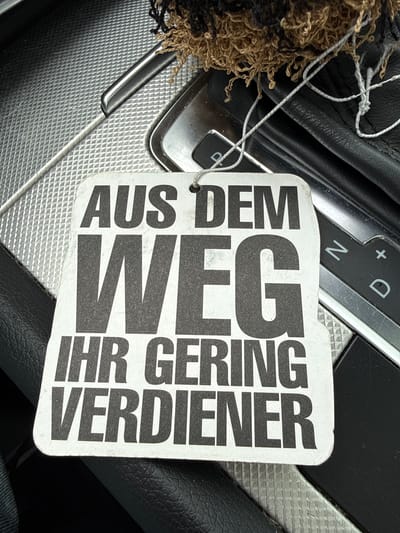 This is a close-up, overhead shot of a car's center console with an air freshener hanging from it. The air freshener is white with bold black German text. The text reads "AUS DEM WEG IHR GERING VERDIENER". The console appears to be made of textured plastic and metal. A gear shift lever is visible to the right, with markings for "N" (neutral), "D" (drive), and "+/-" (manual mode). A fuzzy brown and black object, possibly a decoration or part of the air freshener's string, is also visible at the top of the image. The lighting suggests it is daytime. The specific location within Vellmar, Germany cannot be determined from the image alone.