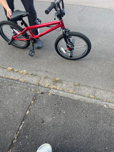 A red HILANO "Redeem" BMX bicycle is positioned on an asphalt surface. The bicycle is in the center of the image, facing slightly to the right. It has black handlebars, a black seat, and black pedals. The front tire appears to be slightly deflated. A person, partially visible from the waist down, is standing to the left of the bicycle, holding the seat with their left hand. They are wearing black pants with white stripes and grey Crocs-style shoes. The background consists of more asphalt, with some cracks and sparse vegetation growing through them, suggesting an outdoor, possibly urban or suburban, environment. The scene does not provide specific landmarks to confirm its location in Wiehl, Germany.