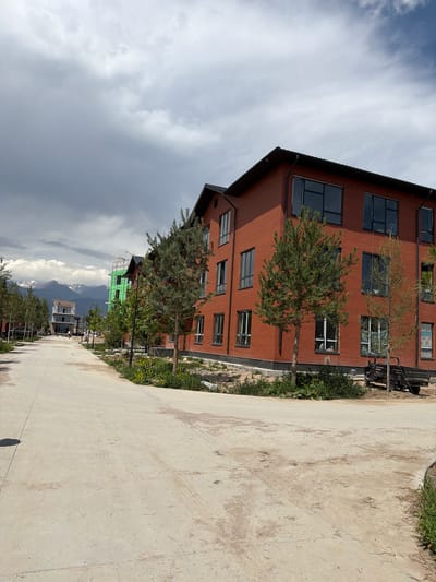 The image depicts a street scene in the city of Kara-Oy, Kyrgyzstan, with a focus on new construction. A paved road curves through the foreground, leading towards a row of red brick buildings. These buildings appear to be multi-story apartment complexes or townhouses, with dark-framed windows. One building in the mid-ground is partially covered in bright green construction sheeting, indicating ongoing work. Further in the distance, more buildings are under construction, including one with a modern, glass-fronted design. In the background, snow-capped mountains rise against a cloudy sky, suggesting a mountainous or sub-alpine setting. The scene is populated by young evergreen trees planted along the roadside and in front of the buildings. A small, dark utility cart is parked on the right side of the road near one of the brick buildings. The overall impression is one of development and expansion in a scenic, potentially suburban or resort area.