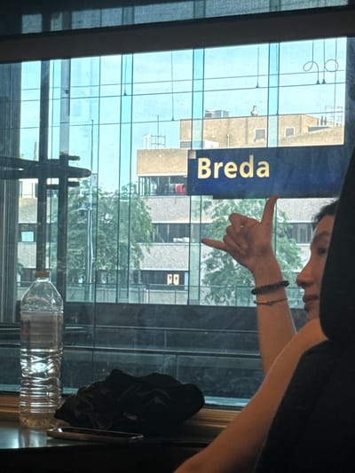 The media file shows a close-up of a person's hand gesturing towards a blue sign with yellow text that reads "Breda". The sign appears to be a directional sign, possibly indicating a train station or a location within a city. The person's hand is positioned in a "shaka" or "hang loose" gesture, with the thumb and pinky finger extended. They are wearing a dark bracelet on their wrist.
The background of the image is a reflection on a glass surface, showing a cityscape with modern buildings and trees. The lighting suggests it is daytime, with bright natural light. The reflection indicates the photo was taken indoors, looking out through a window. The presence of a plastic water bottle in the foreground, partially obscuring the reflection, suggests a casual or travel-related context. The overall mood is relaxed and casual, hinted at by the gesture. The location context provided, Wuustwezel, Belgium, might be relevant to "Breda" as a destination or route from that area.