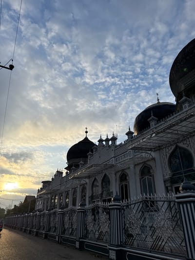 This image captures the At-Taqwa Mosque in Keudekruenggeukueh, Indonesia, at either sunrise or sunset. The sky displays a mix of cirrocumulus and altocumulus clouds, with warmer yellow and orange hues near the horizon on the left, transitioning to cooler blues and greys higher up and to the right. The sun is visible as a bright, diffused orb on the far left, casting an intense, warm glow across the lower clouds and the scene.
The mosque, a prominent structure, occupies the central and right portions of the frame. It features multiple dark, possibly black or very dark blue, domed roofs, some topped with a crescent moon and star finial. The facade of the mosque is predominantly light-colored, likely white or light grey, adorned with intricate architectural details including arched windows, decorative patterns, and multiple pointed spires or finials along the roofline. A multi-paneled, transparent canopy structure extends from the facade over several windows.
A decorative fence, composed of vertical striped posts and intricate geometric metalwork panels, runs along the base of the mosque, separating it from the adjacent road. The road surface appears to be asphalt. On the far left, partially obscured by the fence and the sun's glare, a motorcycle with a rider is visible, moving away from the viewer. Utility poles and overhead power lines are visible on the upper left, extending vertically into the sky. Scattered trees and low-rise structures are discernible in the background behind the fence and road on the left. The overall lighting suggests a low sun angle, with parts of the mosque and road in shadow and highlights catching the details of the facade and fence.