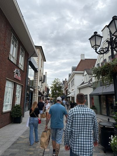 This image captures a bustling street scene in Roermond, Netherlands, likely within the Designer Outlet. The perspective is from behind a group of people walking down a cobblestone path lined with retail establishments. The architecture features a mix of brick and white-washed buildings with red tile roofs. Overhead, a cloudy sky looms.

In the foreground, two men are walking away from the camera. The man on the right wears a blue and white plaid shirt and jeans. The man to his left wears a blue t-shirt and denim shorts. They are walking on the right side of the path.

Further down the path, a woman in flared jeans and a black top walks away from the camera, carrying a white shopping bag in her left hand and a red handbag on her shoulder. She is followed by another woman in a white blouse and shorts, holding a shopping bag. Several other shoppers are visible in the middle and background, some carrying shopping bags, indicating a commercial area.

Visible storefronts include "Swarovski" and what appears to be "Marc O'Polo." The building on the left is a red brick structure with large windows and signage for "Staub." Traditional street lamps with ornate black metalwork and glass lanterns are interspersed along the street. Greenery in planters adds to the atmosphere. The overall scene depicts a lively shopping district.