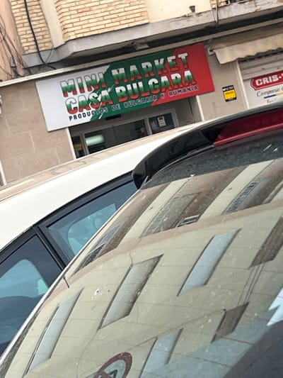 This image depicts a street scene in Ontinyent, Spain. The central focus is a storefront featuring a prominent sign for "MINI MARKET CASA BULGARA". The words "MINI MARKET" and "CASA BULGARA" are rendered in segmented block letters, incorporating white, green, and red, which visually align with the Bulgarian flag. Beneath this, smaller white text on a green background states "PRODUCTOS DE BULGARIA Y RUMANIA" (Products of Bulgaria and Romania). The overall sign is predominantly red with green accents.

Above the storefront, a building constructed with light brown bricks is visible. To the right of the Mini Market, an adjacent establishment has a partially visible sign displaying "CRIS-" and "Companie", with a beige awning retracted above it. A small yellow rectangular sign is affixed to the wall between the two businesses.

In the foreground, the view is partially obstructed by the rear window and roof of a white vehicle. The car's rear window is visibly dirty and reflects the facade of a building opposite, showing multiple rectangular window patterns. A red spoiler or third brake light is integrated into the top edge of the car's rear. A circular reflection, possibly of a road sign, is faintly visible in the lower right portion of the car's rear window.