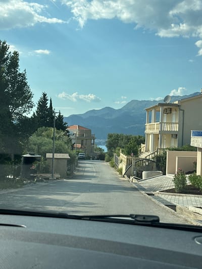 This media file captures a view from inside a vehicle, looking out onto a street lined with buildings and lush greenery. The scene is dominated by a large, imposing mountain range in the background, its peaks shrouded in a light haze. The sky above is a vibrant blue, dotted with scattered white clouds, suggesting a clear and pleasant day.
On the right side of the frame, a paved road stretches into the distance, casting long shadows from unseen trees, indicating it is likely late afternoon or early morning. Adjacent to the road, there are several multi-story buildings, some with balconies and decorative elements, suggesting a residential or vacation area. Dense green foliage, including trees and bushes, flourishes between the buildings and along the roadside, adding a natural element to the urban landscape. A satellite dish is visible on one of the buildings.
There are no people clearly visible in the frame, and the focus is on the static elements of the environment. The overall impression is one of a quiet, sunny day in a picturesque location, possibly a coastal town given the presence of mountains and what appears to be distant water in the far background, though this is not clearly discernible. The media file is oriented vertically.