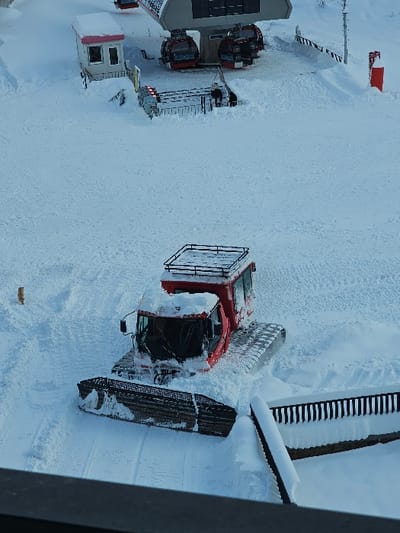 The image captures a snow-covered ski resort area in Danisparauli, Georgia, under winter conditions. In the midground, a red snow groomer, featuring tracks and a front plow blade, is stationary on groomed snow, partially covered by fresh accumulation. To its immediate right, a black vertical-slat fence is partially buried in snow. In the background, an elevated gray ski lift station holds several red gondola cabins, which appear static at a loading platform. A small, snow-covered building with a pink roof and windows is situated to the left of the lift station. Two indistinct human figures are visible near the gondola platform. A red cylindrical marker stands upright in the snow to the far right of the scene. The environment is uniformly covered in snow, comprising both groomed and naturally accumulated sections.