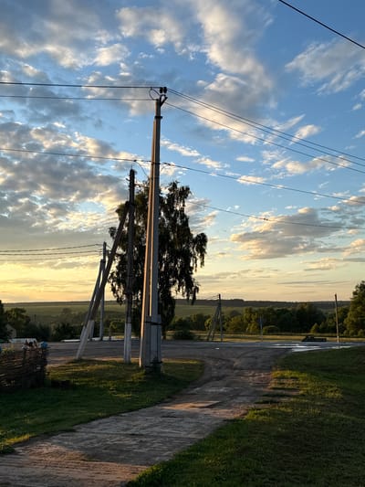 This image depicts a rural landscape in Petrovskoye, Russia, at sunset. The scene is dominated by a dirt road running diagonally from the lower right to the upper left, flanked by lush green grass. In the distance, a treeline marks the horizon, and a vast expanse of sky filled with dramatic, golden-hued clouds stretches above.
Prominently featured are several utility poles with overhead wires, adding a touch of man-made infrastructure to the natural setting. The warm glow of the setting sun casts long shadows and illuminates the clouds with shades of orange, yellow, and pink, suggesting a peaceful and serene atmosphere. The overall impression is one of tranquility and the quiet beauty of the countryside at the end of the day. There is no visible text in the image.
