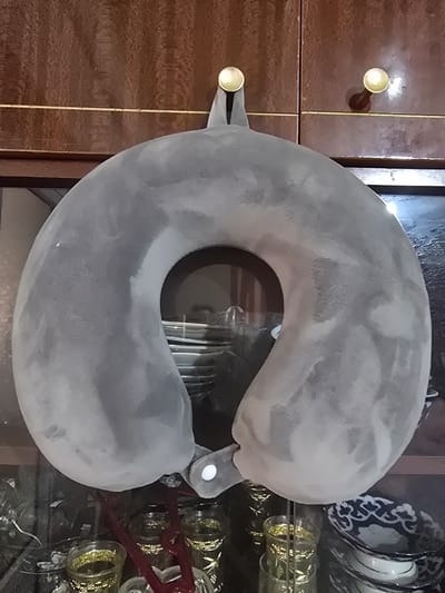 The image shows a gray U-shaped travel pillow hanging by a thin gold string from two golden knobs on a wooden cabinet. The cabinet has a textured brown wood grain. The pillow has a small white button on its underside, near one of the ends. In the background, through the opening of the pillow and the glass doors of the cabinet, various objects are visible, including what appear to be decorative bowls with blue and white patterns, and several glasses filled with a yellow liquid and illuminated with small lights. The setting is likely a home or a shop, possibly in Karasu, Uzbekistan, given the specific mention of that location.
