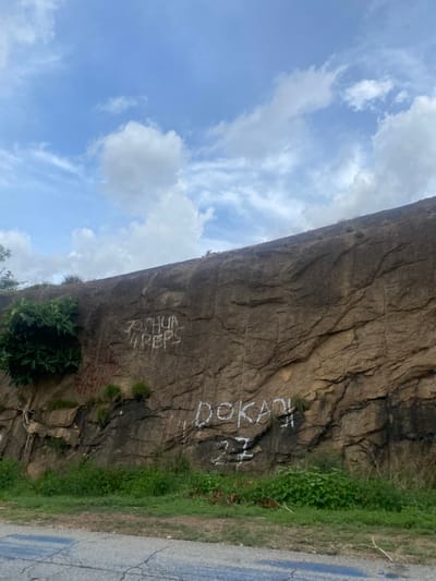 The image shows a rocky cliff face with graffiti on it. The cliff is in a natural setting, with grass and some greenery growing at its base and along the top edge. A paved road is visible on the right side of the image, bordering the grassy area. The sky above is partly cloudy with patches of blue sky and white clouds.
The graffiti on the rock face appears to be in white paint and includes several words or tags. One prominent tag reads "DOKAN Z." Below that, another tag appears to be "TOBIA" or "TORIA" followed by "REP". There are also some red markings on the rock face, though they are less legible.
The lighting suggests it is daytime, likely late morning or early afternoon, given the brightness of the sky and the way the light falls on the rock. The weather appears to be clear and pleasant, with sunshine and scattered clouds. The location context of Rimin Ziem, Nigeria, provides a geographical setting for this scene. No people are visible in the image. The overall impression is a rugged natural landscape marked by human intervention through graffiti.