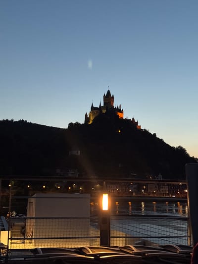 The image depicts Cochem Castle, a prominent landmark in Cochem, Germany, silhouetted against a twilight sky. The castle, illuminated by warm yellow lights, sits atop a dark, tree-covered hill. Below the castle, the lights of the town are visible, reflecting on the surface of a body of water, likely the Moselle River. In the foreground, part of a deck or balcony is visible, with lounge chairs and a light fixture. The overall scene suggests an evening view, possibly from a boat or a riverside vantage point. There are no people or animals visible in the image.