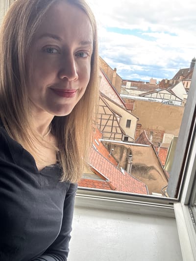 The image shows a medium shot of a Caucasian woman with blonde hair, fair skin, and blue eyes, wearing a dark v-neck shirt and a pearl necklace. She is looking directly at the camera with a slight smile. Behind her, a window reveals a view of the rooftops of Colmar, France. The architecture suggests a historic European city, with terracotta tiles and half-timbered buildings. The sky is partly cloudy. The lighting appears to be natural, coming from the window.