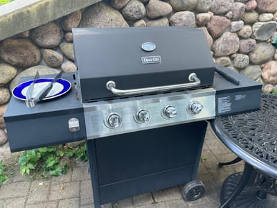 This is a top-down, outdoor shot of a black propane grill, likely in a backyard setting. The grill is positioned on a stone patio, with large, varied-colored stones forming the ground cover. To the left of the grill, a portion of a black, ornate metal table is visible, suggesting a dining or seating area. A small, black wheel, possibly belonging to a grill cart or similar item, rests on the paved area behind the table. Green foliage peeks out from behind the grill and the stones, indicating a natural element in the surroundings.
The grill itself is the primary subject. It features a stainless steel control panel with four knobs, each with markings and a flame indicator. A silver handle spans the front of the grill's main body, below the control panel. The brand name "Dyna-Glo" is visible on the front of the grill, along with a temperature gauge. On the right side of the grill, a white plate with a blue rim is placed, holding a pair of metal tongs, as if ready for or having just finished a meal.
There is no visible indication of the time of day or specific weather conditions, though the lighting suggests it is daytime. The overall scene evokes a casual, domestic outdoor environment, possibly a patio or garden. No people are visible in the image.
Visible text includes: "Dyna-Glo" on the front of the grill, and various instructions and warnings on a label on the side of the grill panel, though the text is too small to be fully deciphered in the provided image.