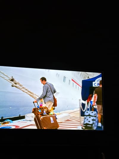 The image shows a television screen displaying a scene depicting a man disembarking from a cruise ship. The man is dressed in a gray jacket and light-colored pants. He is pulling two brown suitcases up a gangplank from the ship. The cruise ship has the word "Carnival" visible on the side. The surrounding environment appears to be a sunny day. There is no indication of this being in the city of College Station, United States of America.
