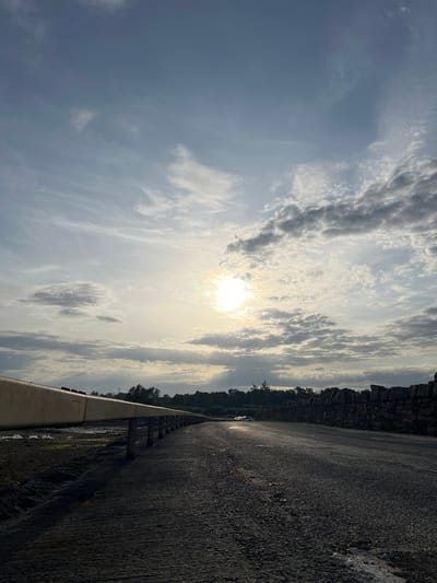 The image is a vertical landscape of a cloudy sky with the sun peeking through, casting a bright glow. The sky is filled with various types of clouds, from wispy cirrus formations to more textured cumulus clouds. In the foreground, a dark asphalt road stretches diagonally across the frame, bordered by a stone wall on the left and a metal guardrail on the right. The road appears wet, suggesting recent rain or morning dew. On the far left, a line of trees and a stone cliff are visible. There is a glimpse of a white car on the road in the distance. The lighting indicates it is either early morning or late afternoon, with the sun low in the sky. The overall mood is serene and somewhat dramatic due to the contrast between the bright sky and the dark road. No text is visible in the image. The scene appears to be a scenic overlook or a road along a cliff face.