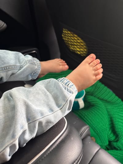 A close-up shot shows the bare feet and lower legs of a child sitting in what appears to be a car. The child is wearing light-wash denim jeans that are rolled up at the ankles, revealing their feet. The left foot is more visible, with five toes clearly discernible. A hint of another bare foot can be seen in the lower left corner.

The background is a mix of car interior elements. To the left, a black mesh pocket is visible, containing a yellow woven object. Behind the child's legs, a bright green knitted fabric is draped, possibly a blanket or clothing. A portion of a dark gray car seat with white stitching is on the right. The lighting suggests it is daytime, with natural light illuminating the scene, although the specific time of day and weather are not discernible. The overall impression is a casual, everyday moment, likely during a car journey. No text is visible in the image. The setting is identified as Somovshchina, Russia.