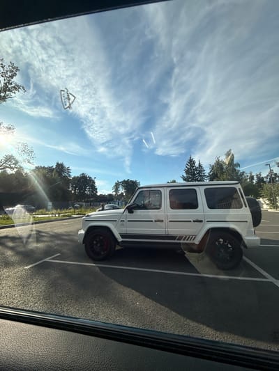 A white Mercedes-Benz G-Class SUV is parked in a lot in Zhukovka, Russia. The sky is bright blue with scattered white clouds. The car is positioned at an angle, facing towards the left of the frame. Its windows are tinted dark, and a spare tire is mounted on the rear of the vehicle. The SUV has black wheels with red brake calipers. The ground is asphalt, marked with white parking lines. Trees and foliage are visible in the background, and a person is standing near a fence in the distance. Sunlight casts shadows across the parking lot. A portion of a vehicle's interior, likely the dashboard and window frame, is visible in the foreground, suggesting the photo was taken from inside another car. A triangle-shaped sticker with text and a picture is affixed to the sky-facing part of the windshield.