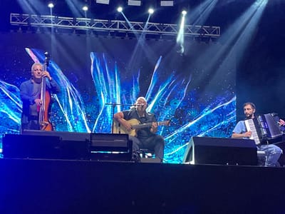 The image displays a musical performance on a stage. Three adult males are positioned across the stage, each playing an instrument. On the left, a male with light grey hair, wearing a blue button-up shirt over a grey t-shirt, stands and plays an upright bass. In the center, a male with a bald head or very short hair, dressed in a dark short-sleeved shirt, is seated and plays an acoustic guitar; a microphone stand is positioned in front of him. On the right, a bearded male, wearing a light blue t-shirt and light-colored ripped jeans, is seated on a white stool and plays a black accordion.

In the immediate foreground of the stage, multiple dark-colored speaker monitors or stage wedges are visible. Above the performers, a metal truss structure with several spotlights directs beams of light downward onto the stage. The background consists of a large LED screen or projection displaying vivid abstract patterns primarily in shades of blue, teal, and purple, featuring swirling lines and circular motifs. The scene indicates a professional concert or event setting, with stage lighting and a prominent visual backdrop. The specific location within the city of Daranak, Armenia, cannot be determined from the visual details present in the image.
