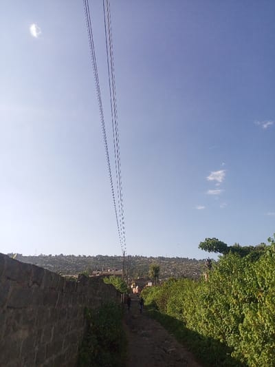 This image depicts an unpaved rural or semi-urban pathway in Kiamaina ward, Kenya, under a clear, bright blue sky with scattered faint white clouds. Multiple parallel overhead utility lines traverse the upper portion of the frame diagonally from upper left to lower right. To the left of the pathway, a dry-stacked stone wall, composed of irregular dark grey rocks, runs along the ground. Lush green foliage grows along the base and crest of this wall. To the right of the pathway, a dense line of vibrant green, leafy bushes and trees forms a natural barrier. Two individuals are observed walking away from the camera along the narrow dirt path, receding into the distance. Both appear to be wearing dark-colored clothing. In the midground, beyond the path and vegetation, a cluster of buildings, some appearing multi-story with light-colored roofs, is visible. A utility pole stands upright among these structures. The background consists of an expansive, undulating hillside covered in sparse vegetation and numerous scattered dwellings, indicating a widespread settlement on the elevated terrain. The scene is illuminated by natural daylight, casting subtle shadows.