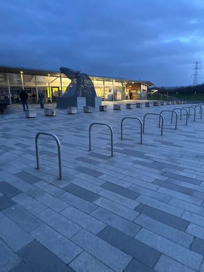 This image depicts an outdoor scene at The Helix, Falkirk, United Kingdom, at dusk or night, under an overcast, dark blue-grey sky.
In the foreground, a paved plaza composed of rectangular grey and darker grey pavers, arranged in a staggered pattern, extends towards the midground. Eight vacant, silver-colored, inverted U-shaped bicycle racks are uniformly spaced and aligned diagonally from the lower left to the middle right of the frame.
The midground features a large, illuminated metal sculpture of a horse's head, consistent with 'The Kelpies' artwork, positioned prominently. The sculpture exhibits an intricate, open mesh construction and is lit from within and below. Directly behind the sculpture is a single-story modern building, identified as a visitor center or retail/food outlet, characterized by extensive glass frontages and a flat roof with external lighting. The interior of the building is brightly illuminated, revealing details through the windows. A series of approximately eight uniform grey concrete bollards are arranged in a linear formation between the paved plaza and the building, with some topped by small yellow conical markers.
Multiple individuals are present: one person in dark clothing walks away from the camera towards the building's left entrance, another stands near a table to the far left of the building, and at least three additional figures are partially visible inside or near the building's entrances.
The background consists of a sloping grassy area behind the building, with a distant electrical transmission tower (pylon) visible against the sky on the far right. A dark treeline or dense vegetation forms the horizon line.