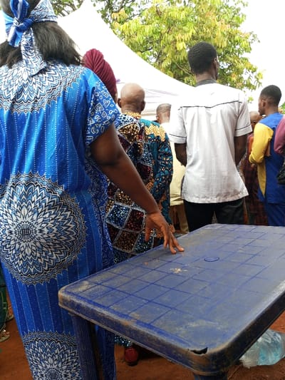 In Oko Town, Nigeria, an outdoor gathering is observed. In the foreground, the back of an individual in a long, blue patterned garment with white intricate designs, including a large mandala motif, is visible. A matching blue and white headwrap is present on the head. The individual's right arm extends forward, with the fingers resting on the surface of a rectangular, worn blue plastic table. The table exhibits a grid pattern and scattered reddish-brown debris.
Beyond this individual, multiple other people are standing. One person directly behind is wearing a multi-colored, diamond-patterned garment in shades of blue, red, gold, and brown, with a bald head and a maroon headwrap. Further back, another person is seen from behind, wearing a white collared shirt with black trim. Additional individuals are partially visible in various attire.
The background features a white canopy or tent, partially obscuring lush green tree foliage. The ground beneath the table appears to be reddish-brown earth. A clear plastic water bottle is positioned on the ground beneath the table.