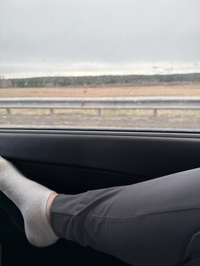 A person wearing grey pants and a white sock sits next to a window inside a vehicle. Outside the window, indistinct scenery is visible, possibly moving. The location within the city of Костенеево, Russia, cannot be determined from the image.