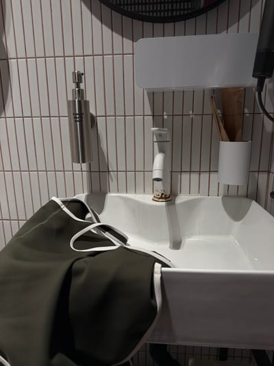 In Kaji-Say, Kyrgyzstan, a close-up shot captures a white bathroom sink with a chrome faucet that shows signs of rust. To the left of the faucet, a stainless steel soap dispenser with Cyrillic and English text is mounted on a white tiled wall. A dark green garment with white trim is draped over the edge of the sink. Above the faucet, a white shelf holds a white cylindrical cup containing wooden toothbrushes and a black hairdryer is visible on the far right. The environment appears to be a bathroom.