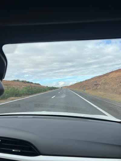 The image displays a straight asphalt highway, observed from the interior of a vehicle through its windshield. The top of the vehicle's dark headliner and the lower portion of its white dashboard, including air vents, are visible in the foreground. The road has two lanes marked by white dashed lines for the median and solid white lines defining the shoulders. Isolated patches of moisture are present on the road surface. On either side, reddish-brown earthen slopes or embankments rise, covered with sparse green and dry vegetation. A blue road marker sign is visible on the right shoulder. The sky is predominantly overcast with white and gray clouds, revealing intermittent patches of blue sky. Distant vehicles are discernible on the road ahead. This scene is located in Ait Ouallal, Morocco.