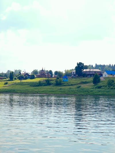 The image depicts a tranquil scene of a riverside village in Ust-Kem, Russia. In the foreground, a body of water with gentle ripples reflects the sky and the distant landscape. The middle ground features a grassy embankment leading up to a cluster of buildings. Among these structures, a prominent wooden church with a dark, tiered roof and a golden dome stands out. Other buildings, including houses and a bright blue gazebo, are visible further along the embankment. The background is filled with lush green trees and a hazy sky with scattered clouds. The overall impression is one of a peaceful rural settlement on a clear day.