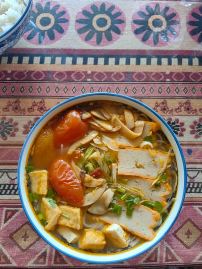 The image shows a bowl of Vietnamese noodle soup on a patterned tablecloth. The soup contains tofu, tomatoes, mushrooms, and what appear to be fish cakes or tofu skins. Chopped green onions garnish the soup. In the upper left corner of the image, there is a bowl of white rice. The photo was taken in Thành phố Hà Giang, Vietnam.
