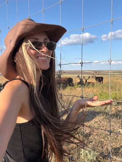 The image captures a woman in a brown wide-brimmed hat and sunglasses, smiling and looking to the right. She is wearing a black tank top and her long brown hair with blonde highlights is blowing in the wind. She is standing behind a wire fence, with her right hand extended towards the fence. In the background, several deer with large antlers are grazing in a grassy field under a blue sky with scattered clouds. The location is identified as Gornyi Balyklei, Russia. The scene appears to be outdoors, possibly a nature reserve or a ranch.