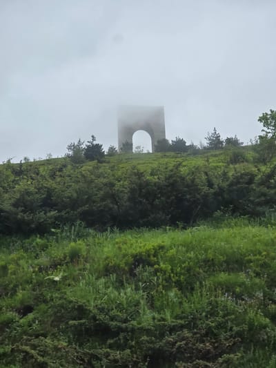 The image depicts a monumental arch structure partially enveloped in dense fog, situated atop a verdant hillside under an overcast sky, near Karnare, Bulgaria. The sky is uniformly gray and heavily clouded, with low-lying fog obscuring the upper portions of the arch, creating a diffused atmospheric effect.
The foreground and midground are dominated by a lush, green landscape consisting of dense vegetation. The lower slopes feature a mix of grasses and smaller herbaceous plants, with hints of purple or blue wildflowers interspersed. Moving upwards, the hillside becomes densely covered with a variety of shrubs and small trees, including coniferous species resembling pines or junipers. These plants exhibit various shades of green, indicating healthy growth.
The central subject is a substantial arch, likely constructed from light-colored stone or concrete. Only its lower and middle sections are clearly visible, including its distinct semicircular archway. The top of the structure is diffused and rendered indistinct by the surrounding fog, blending with the sky. The arch stands prominently on the crest of the hill, surrounded by the aforementioned trees and bushes. No individuals, animals, or explicit human activity are present in the frame. The overall lighting is soft and even, characteristic of an overcast and foggy day.