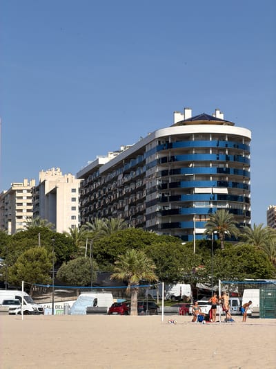 The image shows a group of people playing beach volleyball on a sandy court. The court is lined with a net and poles. In the background, there is a modern, curved building with many balconies and reflective blue windows. Palm trees and other lush green trees surround the building. Beyond the trees, several white vans and a red car are parked. The sky is a clear, bright blue, suggesting it is a sunny day. The people on the court are dressed in athletic wear, and some are in mid-action, either jumping or preparing to hit the ball. A man is standing near a volleyball net on the right side of the frame. The scene appears to be a recreational activity taking place in a coastal urban environment. The text "CAL DEL FIL" is visible on a sign on one of the white vans. The orientation of the image is rotated 90 degrees clockwise.