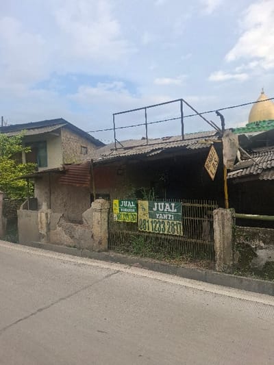 The image shows a partially obscured view of a building located at -6.919, 107.538 in Lagadar, Indonesia. The building appears to be made of concrete blocks and has a corrugated metal roof. It appears to be abandoned or under construction. The building is obscured by a metal fence, overgrown vegetation, and a large sign that reads "JUAL YANTI" and provides a phone number. There is also another smaller sign visible that reads "JUAL TOAKTE" and provides a phone number. The building sits on a hill, and an asphalt road is in the foreground. There is also a partial view of a second building in the background with a similar construction style, and a gold dome of a building is visible in the background behind the second building. The sky is a pale blue with white clouds. No people or vehicles are visible in the scene.