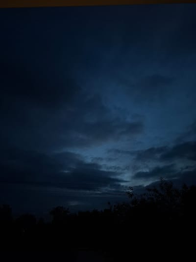 The image captures a dark, cloudy sky with muted blue and gray tones. In the distance, a dark silhouette of trees and foliage forms the horizon line. Thin, dark horizontal lines, possibly power lines, are visible against the sky in the mid-ground. The overall atmosphere is dim and somber, suggesting either dusk or a heavily overcast day. The location is identified as Novlyanka, Russia.