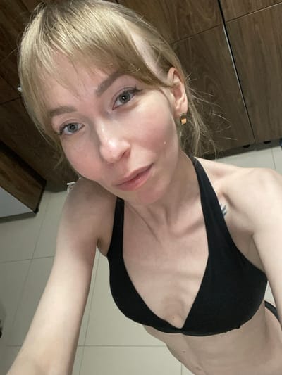 The image is a selfie taken by a woman with long, light blonde hair and light skin. Her head is tilted, and she looks directly into the camera with light blue-grey eyes and a neutral expression. She is wearing a black bra or bikini top. A gold-colored ear cuff is visible on her left ear, nestled within her hair.
A dark, linear tattoo is visible on her upper right shoulder or back area, partially obscured by her arm and the black top. The pose suggests she is angling her body to capture herself from the chest up, with part of her arm and torso visible.
The background indicates an indoor setting, possibly a bedroom or bathroom. On the upper left, there is dark wooden furniture, appearing to be a dresser or wardrobe. The lower and right portions of the background show light-colored tiled surfaces, either a wall or a floor. There is no visible text in the image. The scene is located in Protasy, Russia.