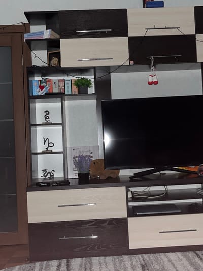 The image depicts an interior scene, likely a living room, featuring a large television mounted on a media console. The console has a mix of dark and light wood finishes, with several shelves and drawers. Various decorative items are visible, including small figurines of dragons, a plant, and a framed picture. A set of miniature boxing gloves hangs from a string on the wall. The overall impression is one of a cluttered but lived-in space. There is no discernible indication of the specific location within the city of Криводановка, Russia.
