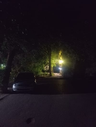 The image shows a street scene at night in Znamensk, Russia. A silver hatchback car is parked on the left side of the frame. The car's license plate is partially visible and appears to read "A243XY 34". A tree with dark foliage is situated to the left of the car. In the background, a bright yellow streetlight illuminates a building and a pathway. The environment appears to be a residential or commercial area with trees and a paved surface. The lighting conditions suggest it is nighttime, with artificial lights being the primary source of illumination.