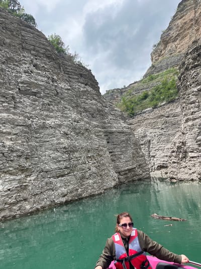 The image shows a person wearing sunglasses and a life vest, possibly in a kayak or small boat, on a body of water with tall, rocky cliffs alongside. The water appears greenish and there's some debris floating on the surface. The cliffs are gray and layered with visible rock formations. There are some patches of vegetation on the cliffs. The sky is overcast. There is no visual information within the image to suggest the location is in the city of Druzhba, Russia.