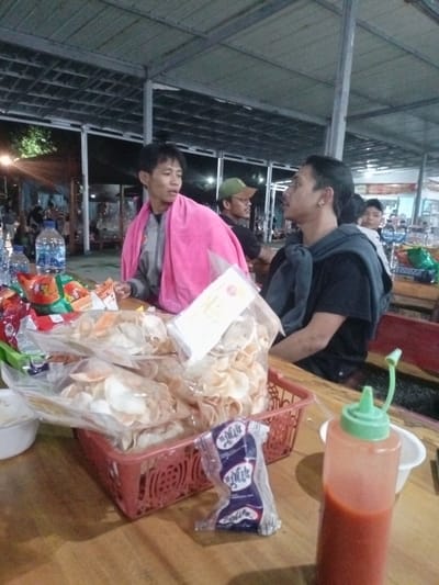 This media file captures an evening scene in Pangalengan, Indonesia, at what appears to be a casual outdoor or semi-outdoor eatery. The setting is dimly lit by artificial lights, indicating it is nighttime, beneath a metallic roof structure.
The foreground features a wooden table laden with various snacks and beverages. Prominently displayed in a red plastic basket are several large clear plastic bags filled with what look like krupuk, a type of Indonesian cracker. To the left, several colorful packaged snacks and two plastic bottles of drinking water are visible. On the right, a tall, clear plastic bottle of red chili sauce with a green cap stands next to a small white bowl. A blue and white packaged snack, with partially legible text including "Mr. P*T*K" and "Susu" (Milk), lies on the table.
Three men are seated at the table. On the left, a man with short dark hair, wearing a gray jacket, has a bright pink towel draped over his shoulders as he looks to his right, engaged in conversation. Beside him and slightly behind, another man wearing a green cap is partially visible. On the right, a man with dark, shoulder-length hair and an earring is seated, wearing a black shirt and a gray top draped over his shoulders, also looking towards the left and participating in the interaction. The background shows other patrons and parts of the eatery, illuminated by ambient lights, suggesting a bustling yet relaxed environment. The overall atmosphere conveys casual dining and social gathering.