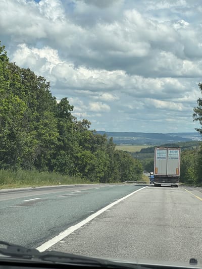 The image shows a view from a vehicle driving on a highway in or near Bavly, Russia. The road is asphalt with white and yellow lane markings. To the left of the road is a grassy embankment lined with trees. To the right, the road curves and goes downhill, with more trees on the right side of the embankment. In the distance, a valley is visible with a town or city and rolling hills under a cloudy sky. A white semi-trailer truck with Cyrillic text on its back is driving ahead on the highway, followed by a blue truck. The text on the white truck appears to be advertising employment, mentioning "ЗАРПЛАТА" (salary) and a phone number. The clouds are white and gray, suggesting a possibly overcast or changeable day.