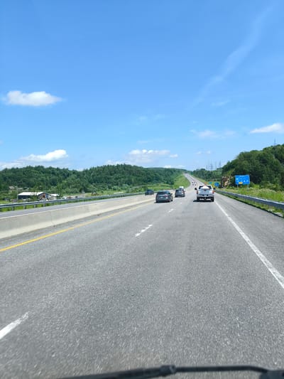 This media file captures a sunny daytime view of a highway in Notre-Dame-de-Bonsecours, Canada. The scene is dominated by a bright blue sky with scattered white clouds. The highway stretches ahead, with several cars driving in the same direction. The road is paved with asphalt and marked with white and yellow lines. On the left side of the highway, there are green rolling hills covered with lush trees, and a building with a grey roof and white siding is visible in the distance. A metal guardrail separates the highway from the grassy shoulder. On the right side, the terrain also features dense green foliage, with tall electrical transmission towers visible further down the road. A blue road sign with white text is present on the right shoulder, indicating directions and distances to "Mascouche," "Joliette," and other locations. The overall impression is one of a pleasant summer drive through a rural landscape.