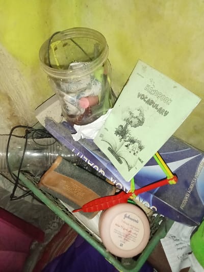 The image displays a collection of miscellaneous household items situated in a corner against a light yellow-green wall in Ciputri, Indonesia. No individuals are visible.
In the upper left, a clear, cylindrical plastic jar containing various small items, including paper scraps and a pink-capped bottle, is positioned. To its right, a worn, light-green booklet titled "HANDBOOK VOCABULARY" with an illustration of figures around an open book rests diagonally. Beneath this booklet, a partially visible blue cardboard box or folder, bearing text such as "QUALITY" and "PRODUKSI AGMATEX INDONESIA," is present.
Tangled dark electrical wires are visible among the objects. A large, empty clear plastic bottle lies horizontally below the jar. A rectangular item with a brown base and dark grey textured top is also present. A red plastic object, resembling a stylized chili pepper, connected to a multi-colored (yellow, green, red) elastic or string component, is centrally located. Next to it, a light pink circular tub of "Johnson's 24 hour moisture soft cream" with English and Arabic text is visible. These items are largely contained within or resting on the edge of a green plastic basket or tray. A partially visible white paper with handwritten notes in Indonesian and a segment of a fuzzy green object are at the bottom right. The scene is static, with no discernible actions or interactions occurring.