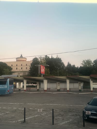 The image depicts a view of the bus station in Lublin, Poland. In the background, a large, beige building with a cylindrical tower on top, resembling a castle or historical fortification, is visible. Trees surround the building and extend towards the bus station. The bus station itself has a covered waiting area supported by pillars. A blue bus is parked on the left side of the frame, with some Cyrillic text on its side. A black Kia car is parked on the right side of the frame, with its license plate visible as "WE 8H715". The ground is a dirt or gravel surface with white painted lines, and there are some bollards and chains. A red KFC billboard with "500" is also visible. The sky is clear and pale blue, suggesting it is either early morning or late afternoon. There are no people clearly visible in the scene.