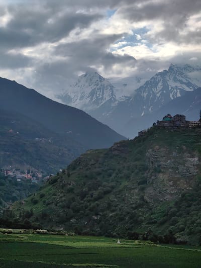 The image shows a mountainous landscape with a village nestled on a hillside in Kardang, India. In the foreground, there are green fields with some white flowers. A small white stupa is visible in the fields. A steep, rocky, and vegetated hillside dominates the middle ground, with a village of multi-story buildings with colorful roofs situated on its crest. Further in the background, more rolling hills are visible, leading to snow-capped mountains under a cloudy sky. The overall scene is one of natural beauty and a rural settlement in a mountainous region.